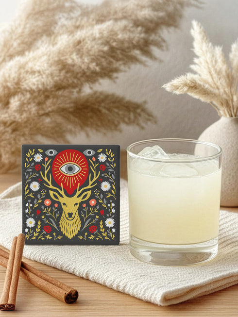 Square ceramic coaster, 4x4 inches, with cork backing. Charcoal tile showcases a golden stag head with antlers, surrounded by florals and an all-seeing eye in red and gold. Smooth printed surface; bold folk-art style. Witchy occult deer coaster for dark academia décor, bar cart, or living room. Square ceramic drink coaster with cork base, stag and eye design.